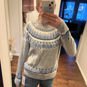 Grå stickad tröja med blått mönster - Säljer en mysig grå stickad tröja med blått och vitt mönster runt halsen och nedtill. Tröjan har rund halsringning och långa ärmar med ribbade muddar. Perfekt för kyliga dagar eller en myskväll🥰🥰