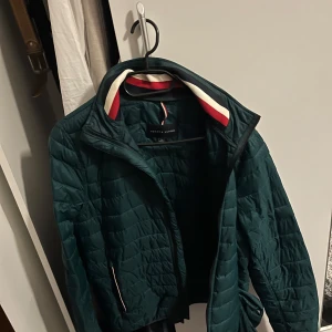 Mörkgrön quiltad jacka från Tommy Hilfiger - Snygg mörkgrön quiltad jacka från Tommy Hilfiger med dragkedja och ståkrage. Fodret har röda och vita detaljer vid kragen. Jackan har långa ärmar och är perfekt för kyligare dagar. Den kommer även med en påse man kan lägga jackan i ifall vädret tillåter.