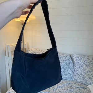 Svart axelväska i syntetmaterial - En stilren svart axelväska med enkel design och dragkedja upptill. Väskan har en mjuk, vadderad axelrem. Perfekt storlek för att bära med sig det viktigaste🌸