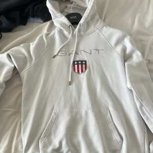 Vit hoodie från GANT - Säljer en stilren vit hoodie från GANT med broderad logga och emblem på bröstet. Den har en klassisk känguruficka och justerbar huva med snören. Perfekt för en avslappnad look.