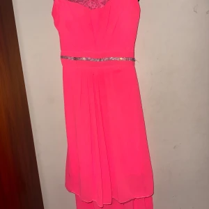 Rosa klänning med spetsdetaljer - Snygg rosa klänning med spets upptill och glittrigt bälte i midjan. Perfekt för festliga tillfällen. Axelbandslös design med en elegant fall.