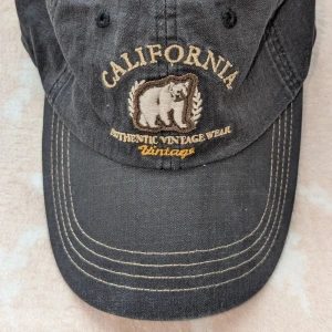 Svart keps med broderad björn - Snygg svart keps med broderad björn och texten 'California' i beige. Kepsen har en vintage-look och är tillverkad av bomull. Perfekt för en avslappnad stil. 100% Bomull. Kepsen har aldrig använts