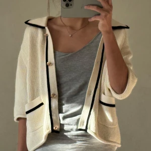 Vit cardigan jacka med svarta detaljer - Snygg vit cardigan jacka med svarta kanter och knappar. Den har kortärmade ärmar och två fickor framtill. Perfekt för en stilren look.