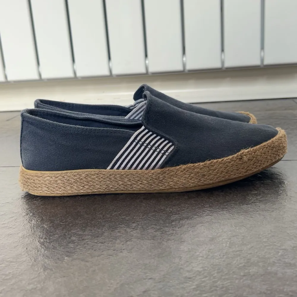 Säljer ett par mörkblå slip-on espadrillos med vit- och svart-randig resår på sidorna. Skorna har en klassisk flätad sula i jute och ovandel i tyg. Perfekta för en avslappnad stil under varmare dagar.. Kengät.