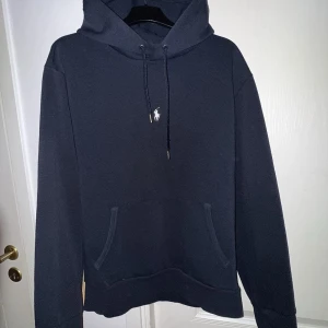 Marinblå hoodie från Ralph Lauren - Säljer en marinblå hoodie från Ralph Lauren med klassisk huva och dragsko. Tröjan har en liten broderad logga på bröstet och en stor magficka framtill. Perfekt för en avslappnad stil.