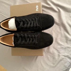  Christian Louboutin - Säljer ett par svarta sneakers från Christian Louboutin med klassisk röd sula och snörning. Skorna är i mocka och har en stilren design med rund tå. Perfekta för dig som vill ha något exklusivt och snyggt till vardags.