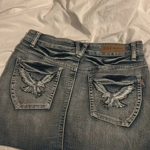 Ljusblå jeanskjol med broderad örn från EDI Fashion - Säljer ett par grå jeans från EDI Fashion med snygga broderade örnar på bakfickorna. Jeansen har klassisk femficksdesign, dragkedja och knapp framtill. Perfekt för dig som vill sticka ut med coola detaljer.