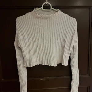 Vit croppad ribbad tröja - Säljer en vit croppad tröja med ribbad struktur och långa ärmar. Tröjan har en hög hals med vågig kant och är perfekt för att skapa en trendig look. Passar till många olika outfits!