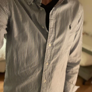 Blåvit randig skjorta från Ralph Lauren - Säljer en klassisk blåvit randig skjorta från Ralph Lauren med knappar framtill och broderad logga på bröstet. Skjortan har lång ärm och normal passform, perfekt till många olika tillfällen.