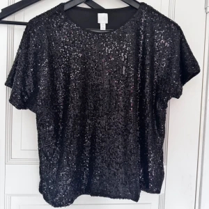 Svart paljettopp från H&M - Säljer en svart glittrig topp från H&M täckt med paljetter. Toppen har rund halsringning och korta ärmar, perfekt för att sticka ut på festen. Snygg och trendig modell som ger en lyxig känsla.