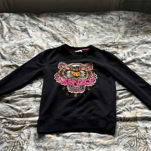 Svart sweatshirt med tigertryck från Kenzo - Cool svart sweatshirt från Kenzo med ikoniskt tigerhuvud och färgglada detaljer framtill. Tröjan har rund halsringning och långa ärmar. Perfekt statement-plagg för dig som vill sticka ut. ⚠️DET STÅR L MEN DEN PASSAR S ⚠️