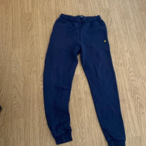 Mörkblå mjukisbyxor från Lyle & Scott Junior - Mörkblå mjukisbyxor från Lyle & Scott Junior med resår i midjan och benslut. Byxorna har en broderad gul logga på ena benet och en bakficka. Perfekta för en avslappnad stil.