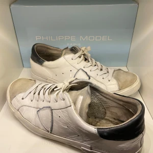 Vita sneakers från Philippe Model - Snygga vita sneakers från Philippe Model med svarta detaljer och snörning. Skorna har en klassisk design med rund tå och är perfekta för en stilren look. Men dom är lite slitna och behöver sig en uppfräschning så som sulan under skon och den har creasningar därav pris…