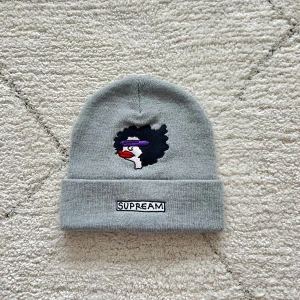 Supreme mössa - Snygg grå mössa från Supreme med en unik broderad design framtill. Mössan har en uppvikt kant och är tillverkad i 100% akryl för en mjuk och bekväm känsla. Perfekt för att hålla värmen med stil.