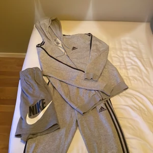 Grå Adidas hoodie och byxor set - Säljer ett grått Adidas set med hoodie och matchande byxor. Setet har svarta ränder längs ärmar och ben samt Adidas-logga på både tröja och byxa. Hoodien har dragkedja och fickor. Perfekt för en sportig look. Du får även med Nike shorts