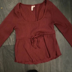 Vinröd ribbad topp från Nelly - Säljer en vinröd ribbad topp från Nelly med lång ärm och knytband i midjan. Perfekt för en stilren look. 💃