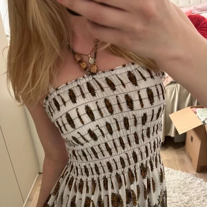 Vit mönstrad sommarklänning med smock - Säljer en vit axelbandsklänning med smock upptill och mönster i brunt, gult och blått. Klänningen har en avslappnad passform och är knälång med elastisk överdel. Perfekt för varma dagar och har ett bohemiskt mönster. Passar storlek S & M  ☺️