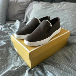 Bruna slip-on sneakers från Michael Kors - Snygga bruna slip-on sneakers från Michael Kors med diskret logomönster och vit platt sula. Skorna har guldfärgade detaljer bak och är perfekta för en stilren vardagslook. Klassisk design med rund tå och bekväm passform. Dem är helt nya oanvända och ordinarie pris är 1695kr den är fortfarande i kartong och plast