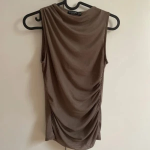 Brun ärmlös topp med drapering - Säljer en brun ärmlös topp från Stradivarius med snygg drapering på sidan. Toppen har en hög hals och är tillverkad i ett mjukt, stretchigt material som sitter tight på kroppen. Funkar som xs/s