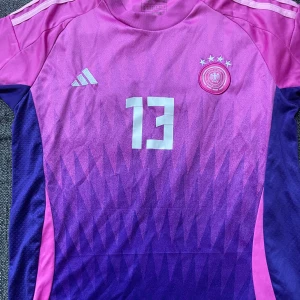  Tysklands Rosa och lila fotbollströja från Adidas - Säljer en snygg fotbollströja från Adidas i rosa och lila med nummer 13 och texten 'Müller' på ryggen. Tröjan har korta ärmar och ett coolt mönster framtill. Perfekt för fotbollsfans som vill sticka ut på planen!