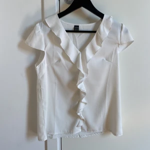 Vit blus från Shein - Säljer en stilren vit blus från Shein med volangdetaljer längs den v-ringade halsen. Blusen har korta ärmar och är perfekt för en elegant look. Passar bra till både jeans och kjol.