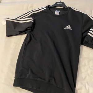 Adidas tröja svart - Säljer en svart kortärmad tröja från Adidas knappt använd  med klassiska vita ränder på axlarna och broderad logga på bröstet. Tröjan har rund halsringning och är gjord i mjukt material, perfekt för en sportig look.