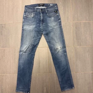 Blå Replay jeansbyxor med slitning - Säljer ett par blå jeansbyxor från Replay med klassisk femficksdesign och lätt slitning på ena benet. Byxorna har normal passform och raka ben. Snygga detaljer på bakfickan och Replay-logga vid fickan fram.