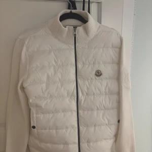 Vit Cardigan från Moncler - Snygg vit quiltad jacka från Moncler med dragkedja och ribbad krage. Jackan har långa ärmar och en stilren design med Moncler-logga på bröstet. Perfekt för kyligare dagar.