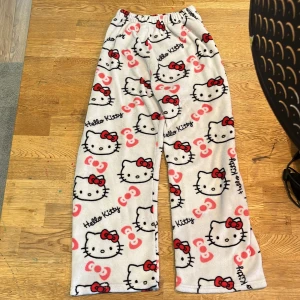 Hello Kitty fleecebyxor - Mysiga vita fleecebyxor med Hello Kitty-motiv och röd/rosa detaljer. Byxorna har resår i midjan och utsvängda ben, perfekta för att chilla hemma eller som pyjamasbyxor.