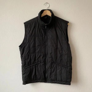 Svart quiltad väst från J.Lindeberg - Säljer en svart quiltad dunväst från J.Lindeberg med dragkedja framtill och ståkrage. Västen har två snedställda fickor och en stilren design som passar perfekt till lager-på-lager-stil. Modell: Gavin Compact Melton - Artnr: Q208