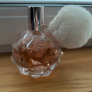 Ariana Grande parfym med vit fluffboll - Säljer en Ariana Grande parfym i en kristallformad flaska med ljusrosa doftvätska och en stor vit fluffboll som dekoration. Flaskan har ett silvrigt lock och en unik, geometrisk design som sticker ut på hyllan.