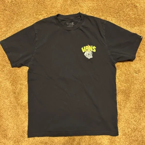 Svart t-shirt med Vans tryck och döskalle - Cool svart t-shirt från Vans med färgglatt tryck av döskalle och skateboard på ryggen samt mindre logga framtill. Perfekt för dig som gillar streetwear och skate-stil. Skön passform och klassisk rund hals. Storlek L barn
