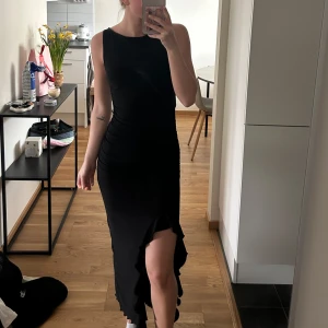 Svart långklänning med volangdetalj - Säljer en elegant svart långklänning med ärmlös design och volangdetalj nertill. Klänningen har en tight passform och öppen rygg, vilket ger en stilren och modern look. Använt en gång