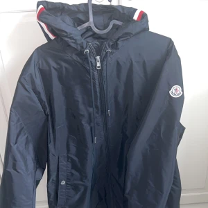 Mörkblå vindjacka från Moncler - Snygg mörkblå vindjacka från Moncler med huva och dragkedja framtill. Jackan har Moncler-logga på ärmen och detaljer i rött och vitt på huvan. Perfekt för blåsiga dagar.