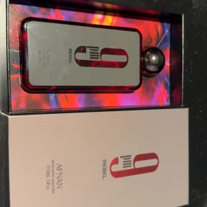 Afnan 9PM Rebel Parfym - Säljer en Afnan 9PM Rebel parfym i en stilren flaska med röd och svart design. Flaskan har en modern rektangulär form med en rund svart kork. Perfekt för den som vill ha en doft med attityd och stil.