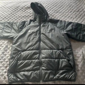 svart pufferjacka från Nike - Säljer en svart pufferjacka från Nike med huva och dragkedja. Jackan har långa ärmar och är perfekt för kyligare väder. Den är designad med Therma-Fit teknologi för extra värme.