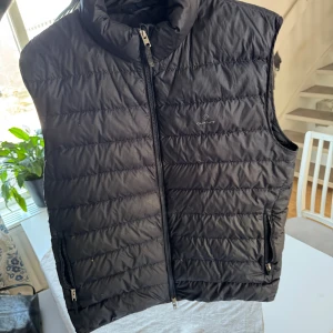 Svart dunväst från GANT - Snygg svart dunväst från GANT med dragkedja och två praktiska fickor framtill. Perfekt för lager-på-lager under kyliga dagar. Västen har en stilren design med diskret logga på bröstet.