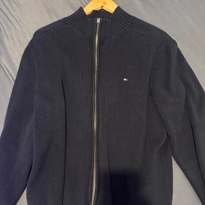 Svart ribbad tröja från Tommy Hilfiger - Snygg svart ribbad tröja från Tommy Hilfiger med dragkedja och diskret logotyp på bröstet. Perfekt för en stilren look.