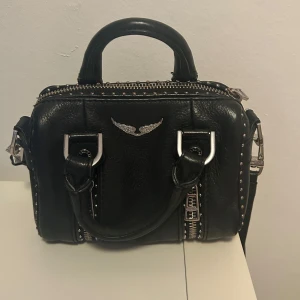 Handväska från Zadig & Voltaire - Snygg svart handväska i skinn från Zadig & Voltaire med coola detaljer. Väskan har en justerbar axelrem med texten 'VOLTAIRE'. Perfekt som festväska men även till vardags. Får plats med dom viktigaste sakerna man behöver ha med sig. Köptes 2022 för 3300kr från Zadig & Voltaires hemsida. Den är i fint skick men inte använd på ett tag så vill att den ska få mer användning hos någon annan.