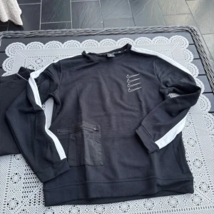 Svart tröja med vita detaljer från Nike - Säljer en svart tröja från Nike med vita ränder längs ärmarna och en liten ficka med dragkedja på framsidan. Tröjan har långa ärmar och flera små Nike-loggor tryckta på bröstet. Perfekt för en sportig look!