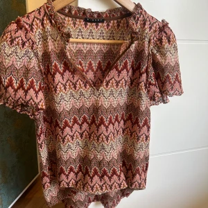 Mönstrad blus med volangdetaljer - Säljer en mönstrad blus i jordnära färger med volangdetaljer vid ärmslut och hals. Blusen har korta ärmar och en v-ringad design. Perfekt för en bohemisk look.