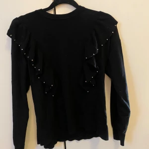 Svart tröja med volangdetaljer från Sfera Basic - Snygg svart tröja från Sfera Basic med volangdetaljer och små pärlor på axlarna. Perfekt för en stilren look. Tröjan har långa ärmar och en normal passform.