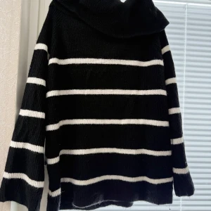 Svart och vit randig stickad tröja - Säljer en mysig svart stickad off shoulder tröja med vita ränder. Tröjan har en hög krage och långa ärmar, perfekt för kyliga dagar.