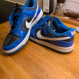 Nike Dunk Low ESS - Säljer ett par Nike Dunk Low sneakers i blått, svart och vitt. Skorna har klassisk låg modell med vit swoosh på sidan och blå snörning. Perfekta för dig som gillar streetstyle och vill sticka ut med färg. Ny pris ligger på 1300