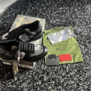 Valentino rockrunners - Snygga Valentino Garavani sneakers i svart och grått med ett coolt camouflage-mönster. Skorna har snörning och en unik design med nitar på sulan. Perfekta för dig som vill ha en stilren men ändå iögonfallande look.
