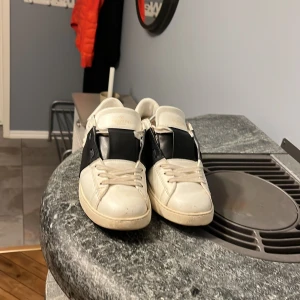 Vit och svart Valentino Garavani sneaker - Säljer ett par vita sneakers från Valentino Garavani med bred svart rem över vristen och diskreta nitar längs sulan. Skorna har rund tå och snörning, samt märkets logga på plösen. Perfekta för dig som gillar stilrena och exklusiva sneakers.priset går att diskutera 