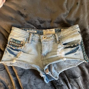 Ljusa jeansshorts med broderade detaljer - Säljer ett par ljusblå jeansshorts med slitningar och broderade mönster på sidorna. Shortsen har nitar längs, perfekta för sommaren! Storlek 36 🌸