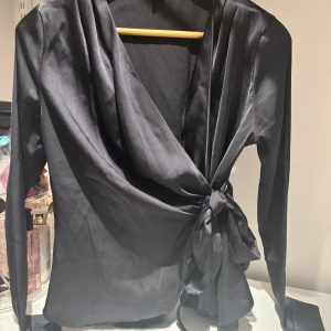 Svart omlottblus med knytband - Snygg svart omlottblus i satin med långa ärmar och knytband i midjan. Blusen har en elegant v-ringning och glansigt material som ger en lyxig känsla. Perfekt att styla till olika outfits.