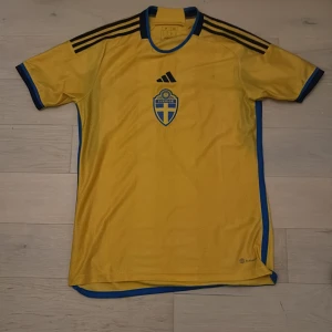 Svenska landslagets fotbollströja från Adidas - Gul kortärmad fotbollströja från Adidas med blå detaljer och svarta ränder på axlarna. Svenska landslagets emblem på bröstet och texten 'Sverige' i nacken. Slim fit-modell med blå kant längs sidor och nederkant.