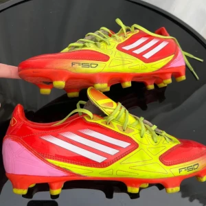 Adidas F50 fotbollsskor i röd och gul - Säljer ett par Adidas F50 fotbollsskor i en riktigt snygg färgkombination av rött, gult och rosa. Skorna har snörning, vita ränder på sidan och är designade för spel på gräs. Perfekta för dig som vill sticka ut på planen!